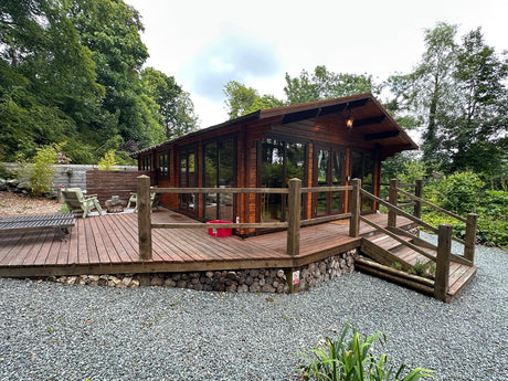 Large timber log cabin with glass doors, spacious wooden deck, outdoor seating, and forest backdrop