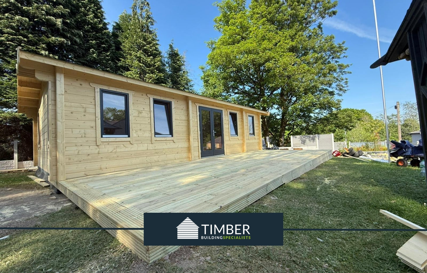 Modern log cabin with large timber deck, set among trees in a garden setting