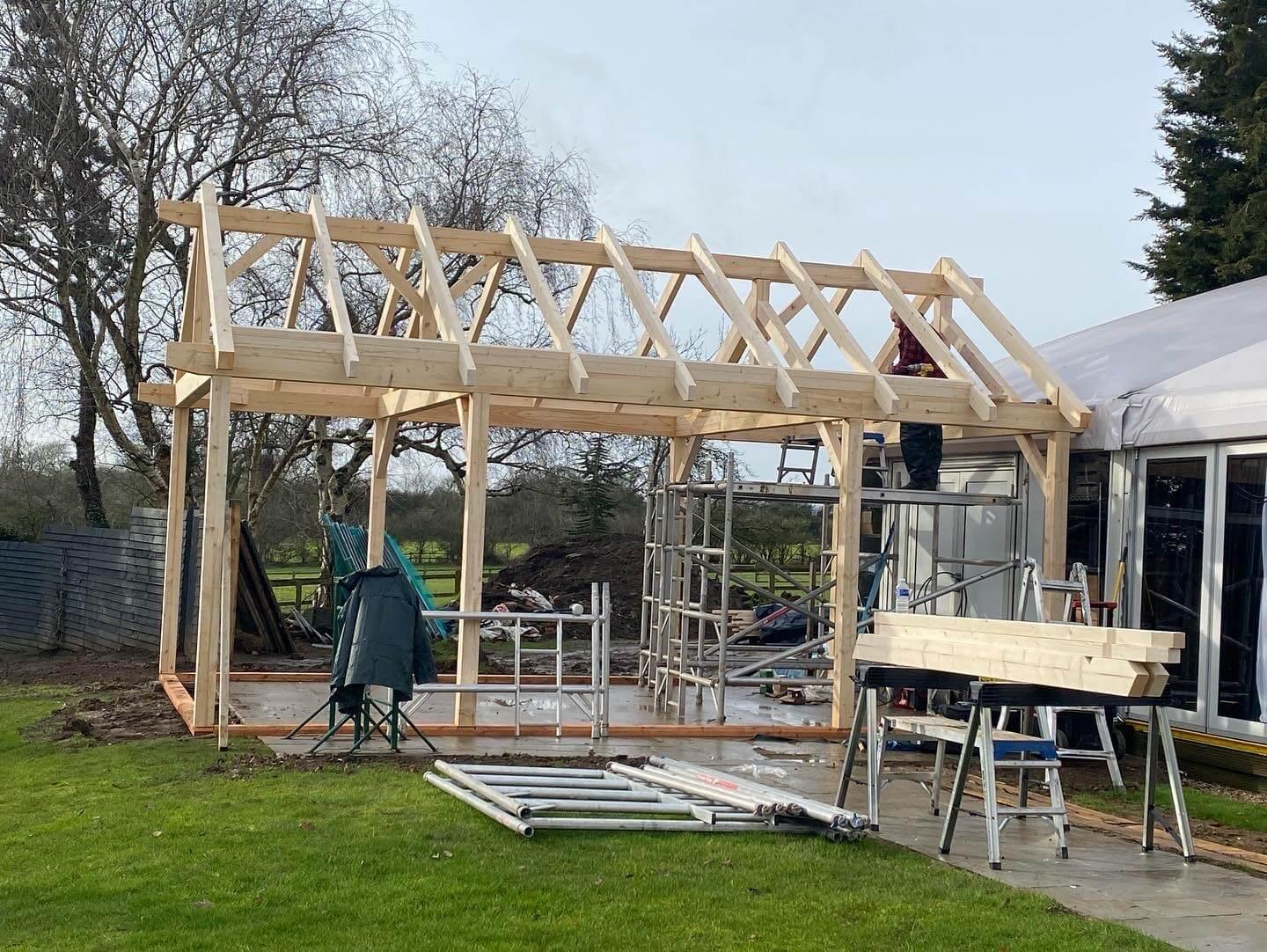 Timber frame log cabin under construction with visible wooden roof beams and outdoor setting