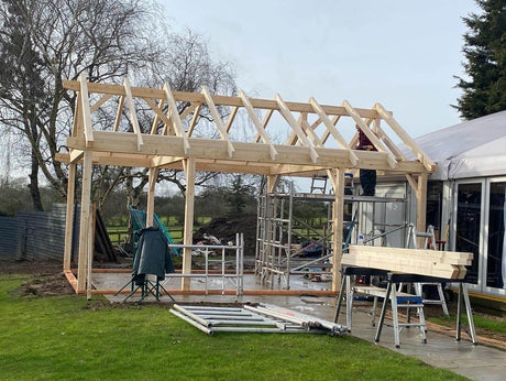 Timber frame log cabin under construction with visible wooden roof beams and outdoor setting