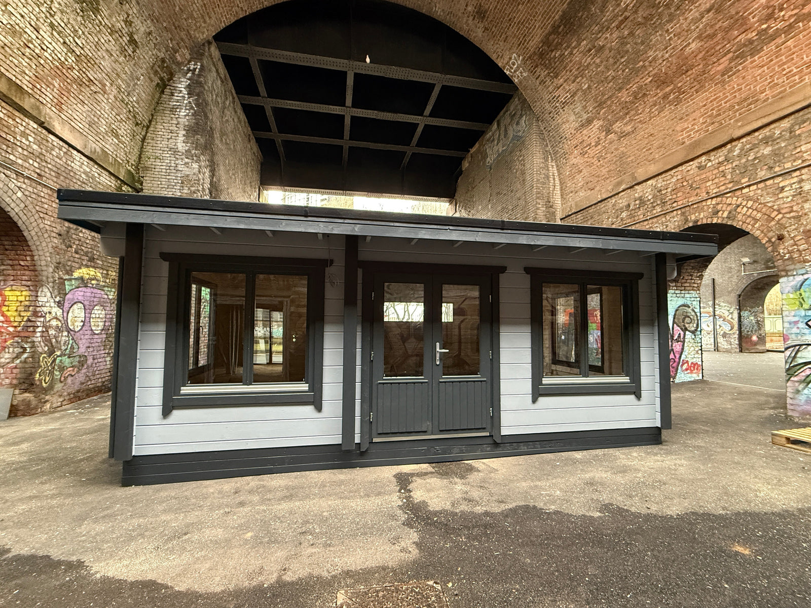 Modern log cabin with double doors and large windows under a brick archway with graffiti walls