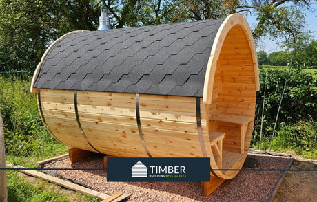 Outdoor barrel sauna made from timber with a shingle roof, set in a garden area.