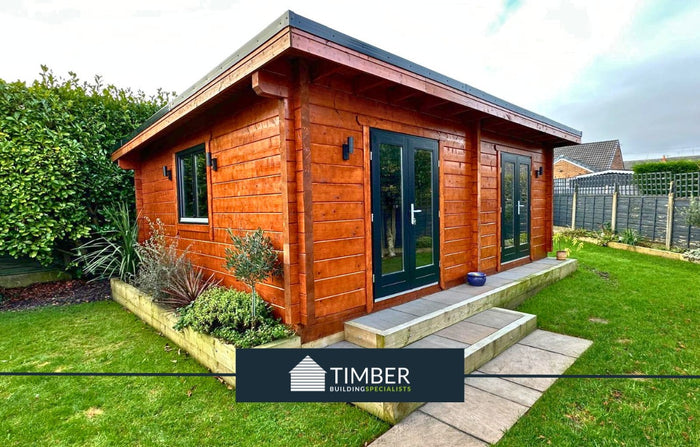 A brown log cabin with black double doors and window, set on a concrete base with a grassy area and shrubbery in the foreground. by the Timber Building Specialists